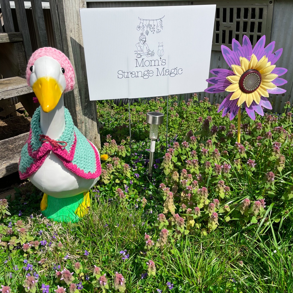 lawn goose with a yard sign in a nature scene with a purple decorative flower.